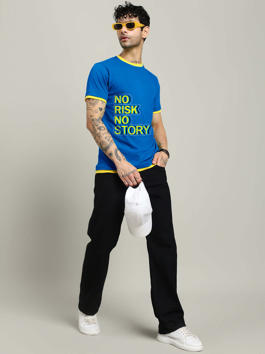 Man wearing NO RISK PRINTED REGULAR FIT T-SHIRT ROYAL BLUE with yellow accents, holding white cap, styled with black pants and sunglasses.