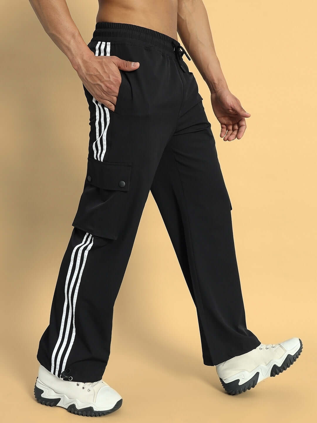 Model wearing NOVA Relaxed Fit Cargo Pants in black with side stripes and white sneakers on a beige background.