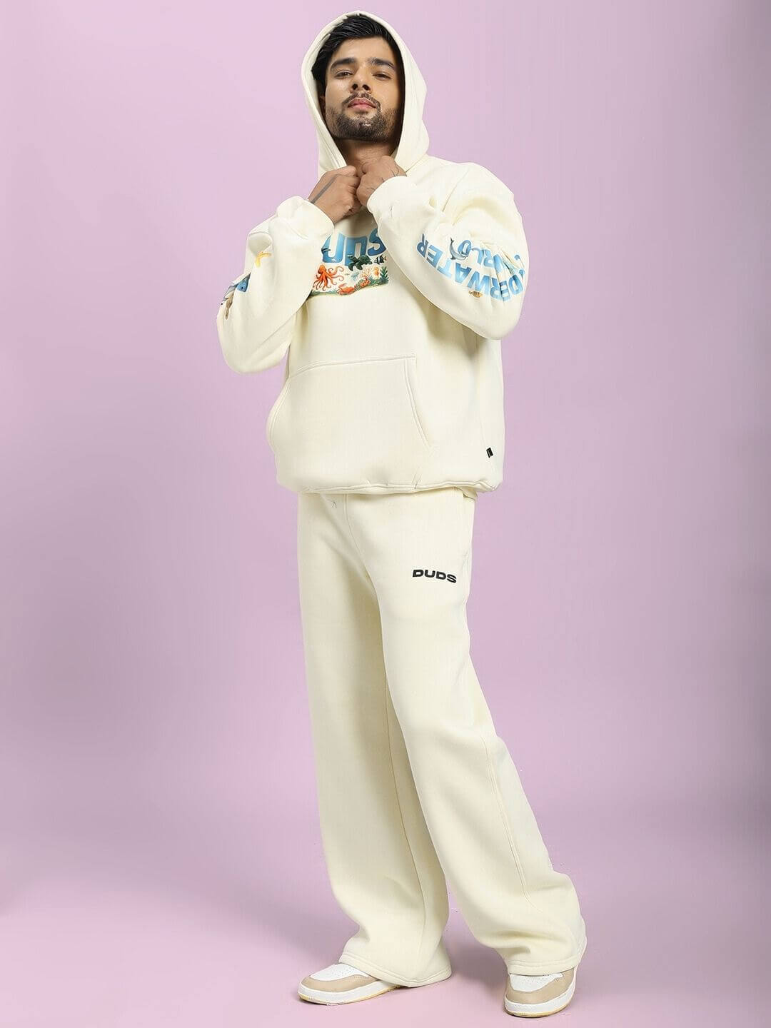 Person wearing OCEAN BUDDIES FLEECE CO-ORD SET OFF-WHITE standing against a pink background, showcasing sea-inspired graphics on hoodie.
