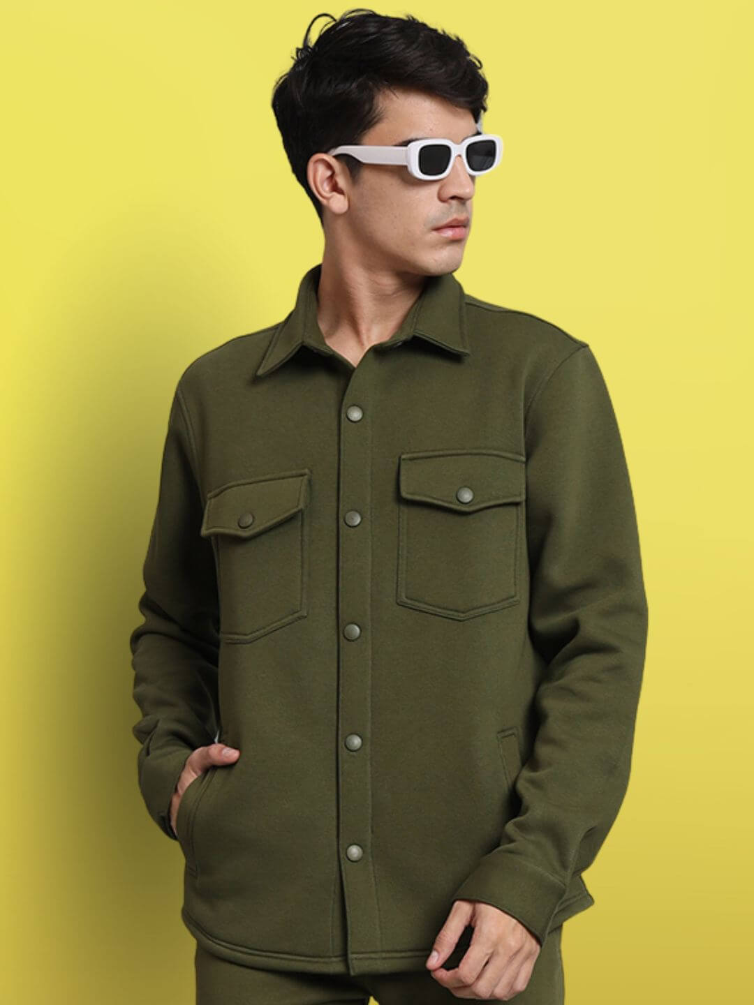 Man wearing olive green relaxed fit full sleeves fleece overshirt and white sunglasses against yellow background.