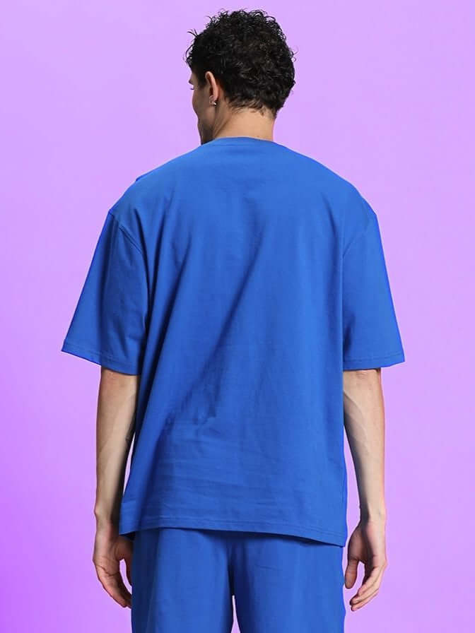 Back view of model wearing OUTLOOK OVER-SIZED T-SHIRT ROYAL BLUE, showcasing the relaxed fit and vibrant color against a purple background.