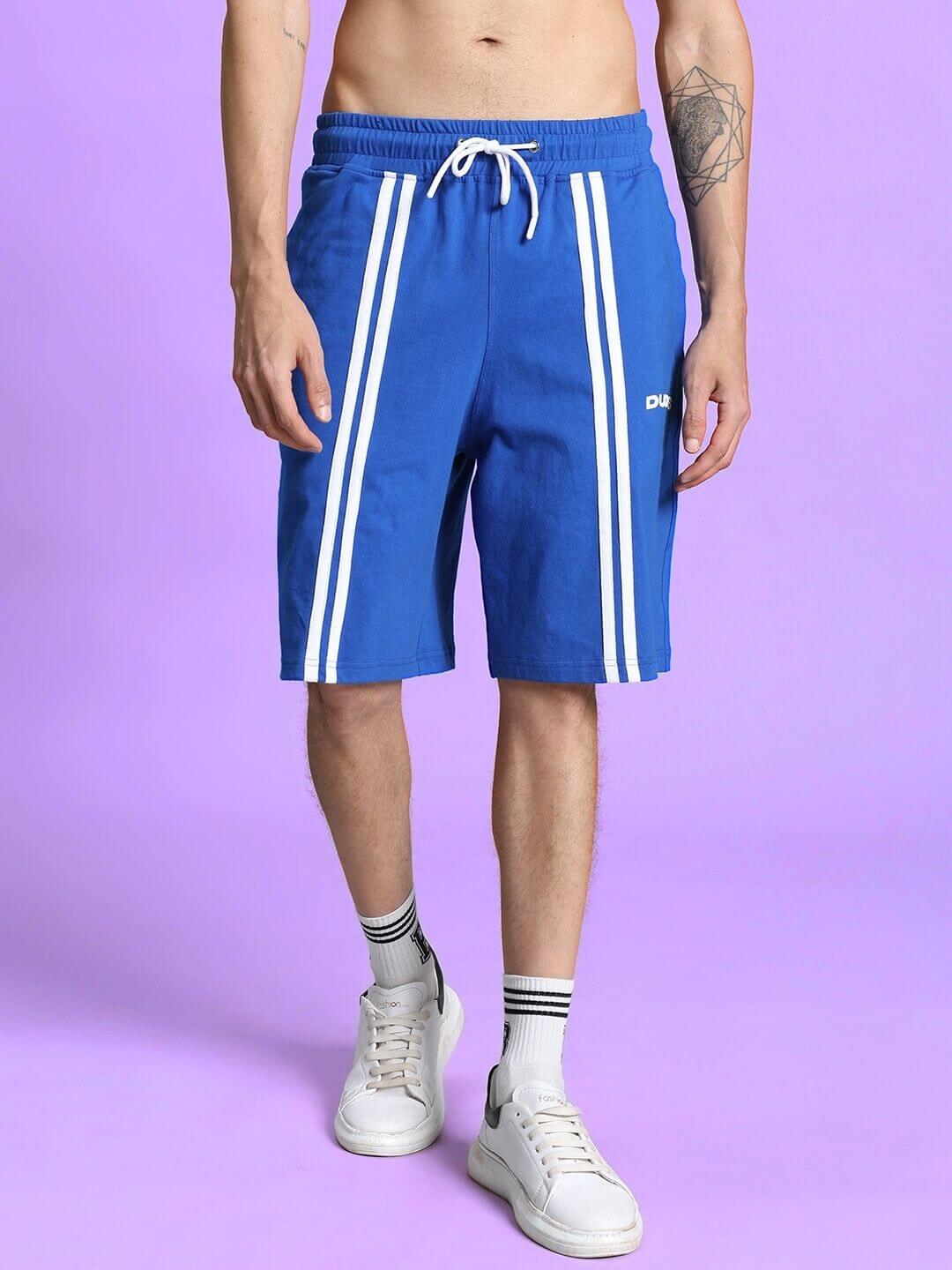 Man wearing OUTLOOK REGULAR FIT SHORTS ROYAL BLUE with white sneakers on purple background.