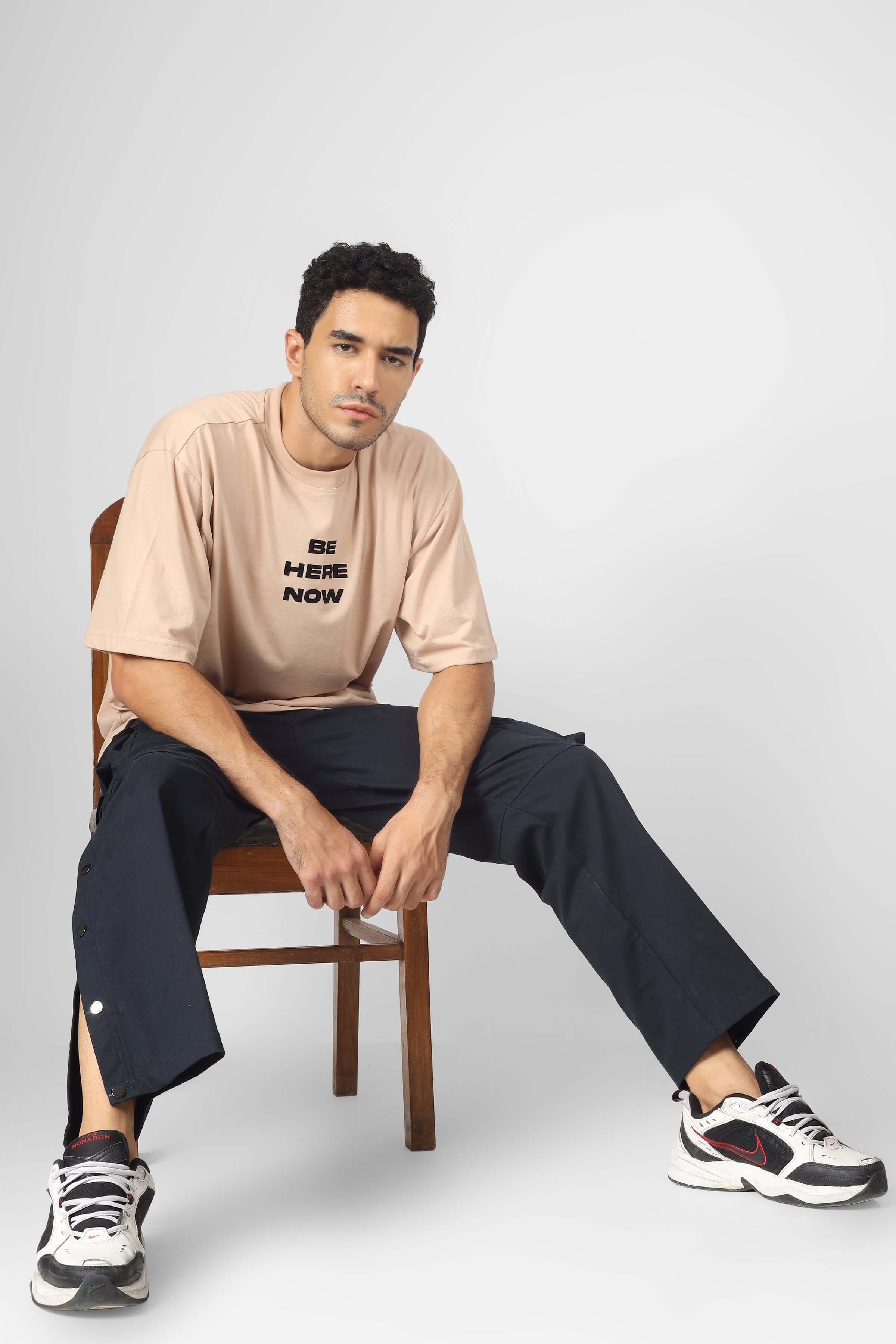 Man wearing Oxford blue cargo pants with six pockets, seated on a chair, styled with casual sneakers and a beige t-shirt.