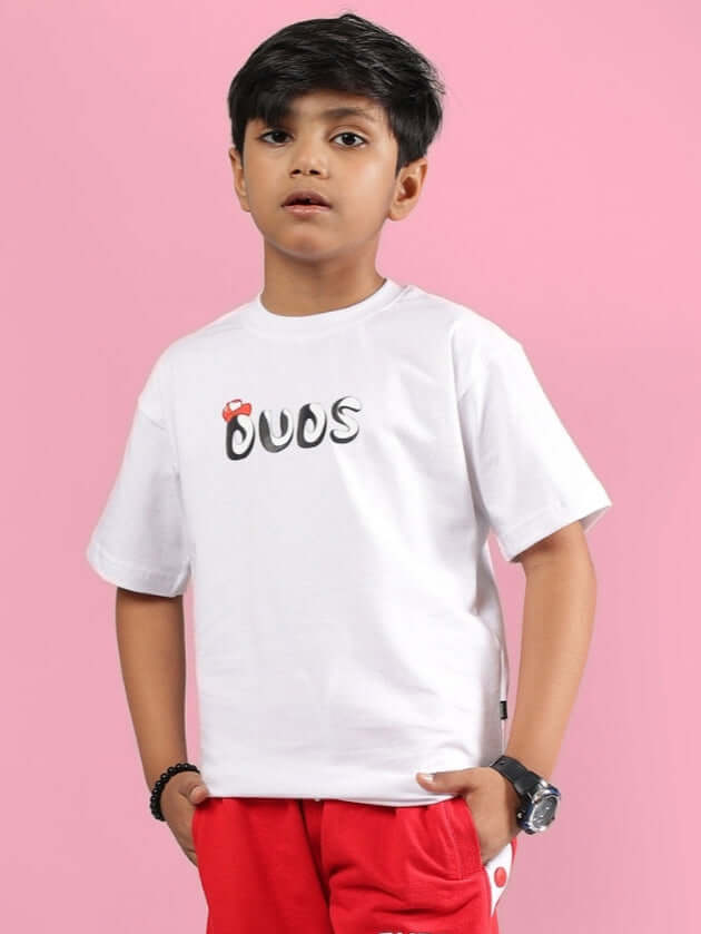 Boy wearing PALOMA T-SHIRT FOR BOYS GIRLS WHITE, styled with red pants, against pink background.