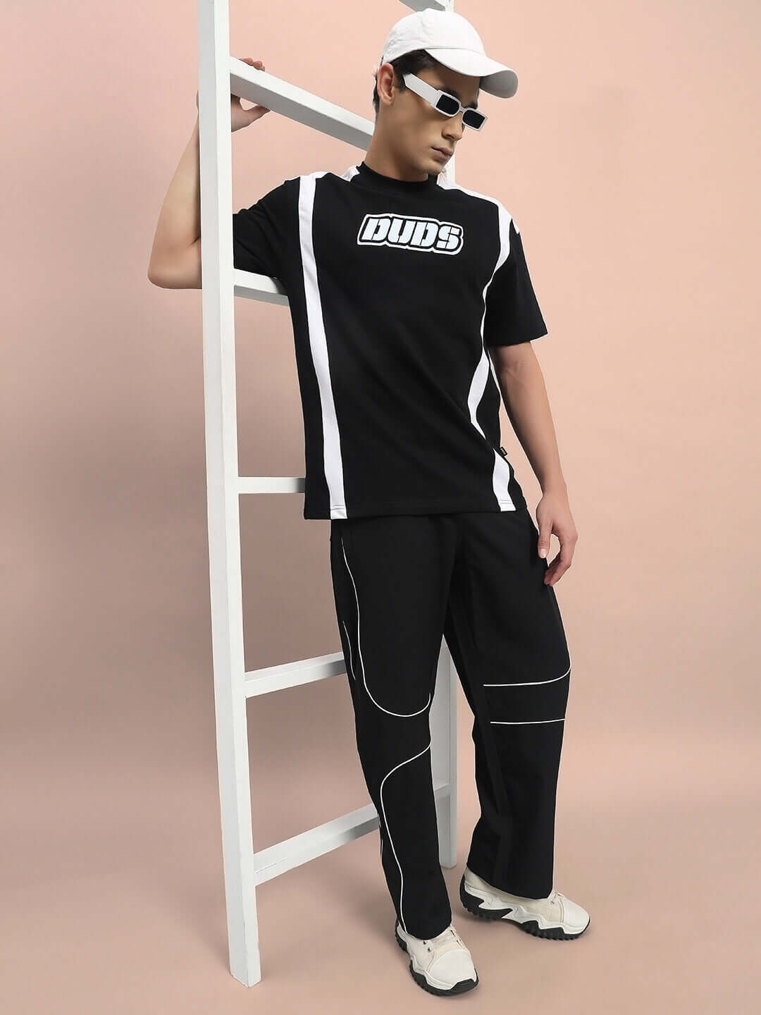 Model wearing DUDS parabolic parachute cargo pants in black with a black and white t-shirt, leaning against a white ladder.