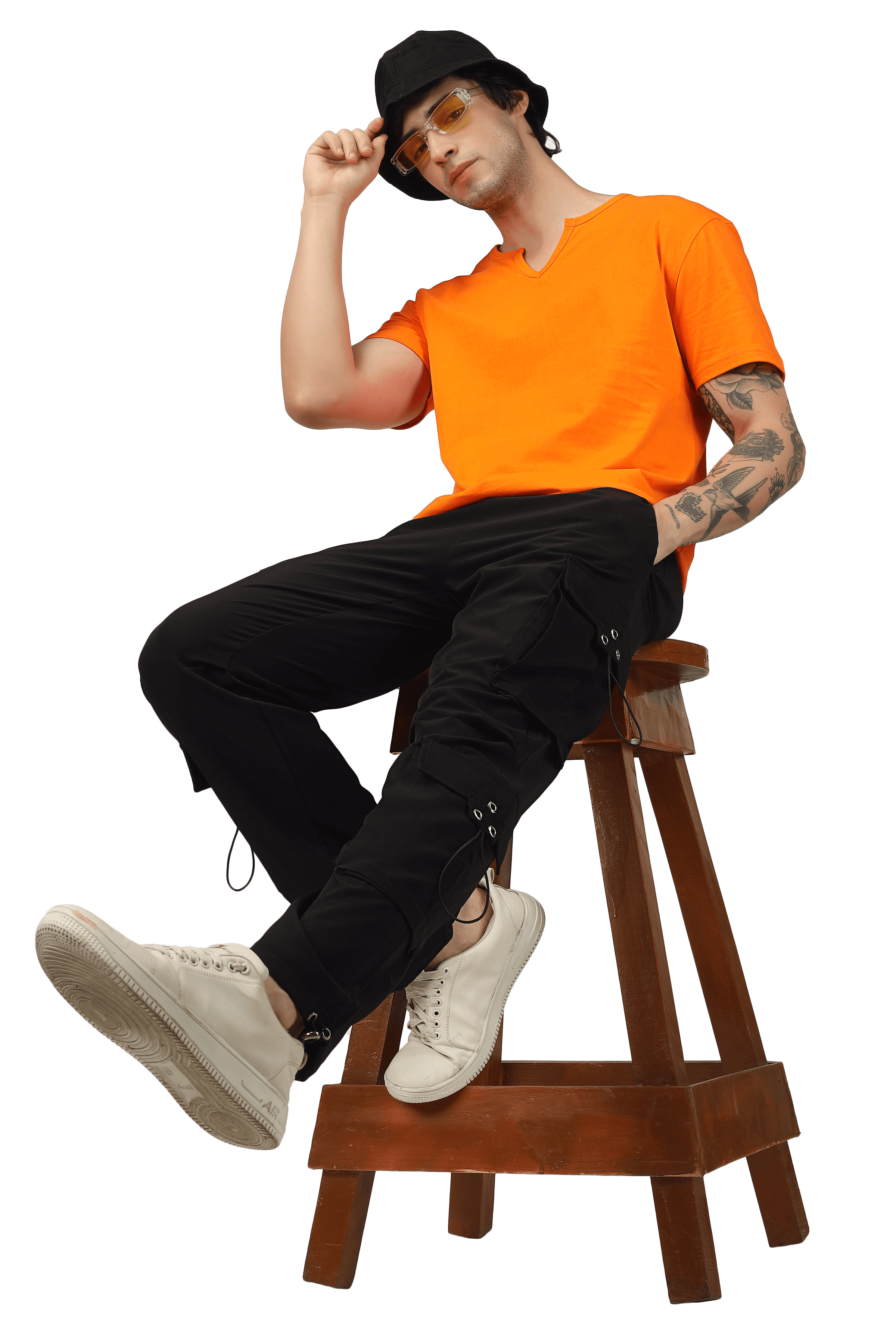 Person wearing black parachute cargo pants with orange t-shirt and white sneakers, seated on a wooden stool, modeling stylish casual wear.