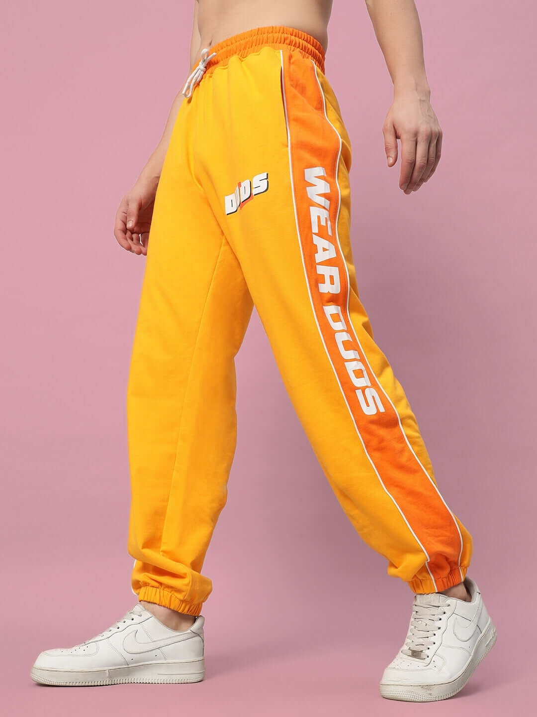 Model wearing Peppy Joggers in yellow-orange by DUDS, highlighted with side stripes and brand logo, paired with white sneakers.