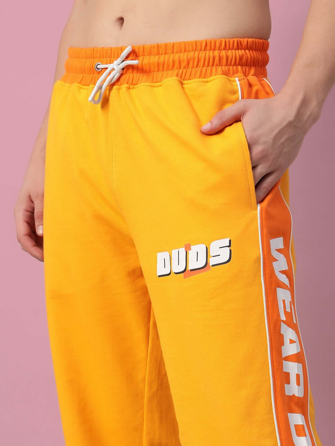 Yellow-orange Peppy Joggers from DUDS featuring logo detail and customizable fit options, against a pink background.