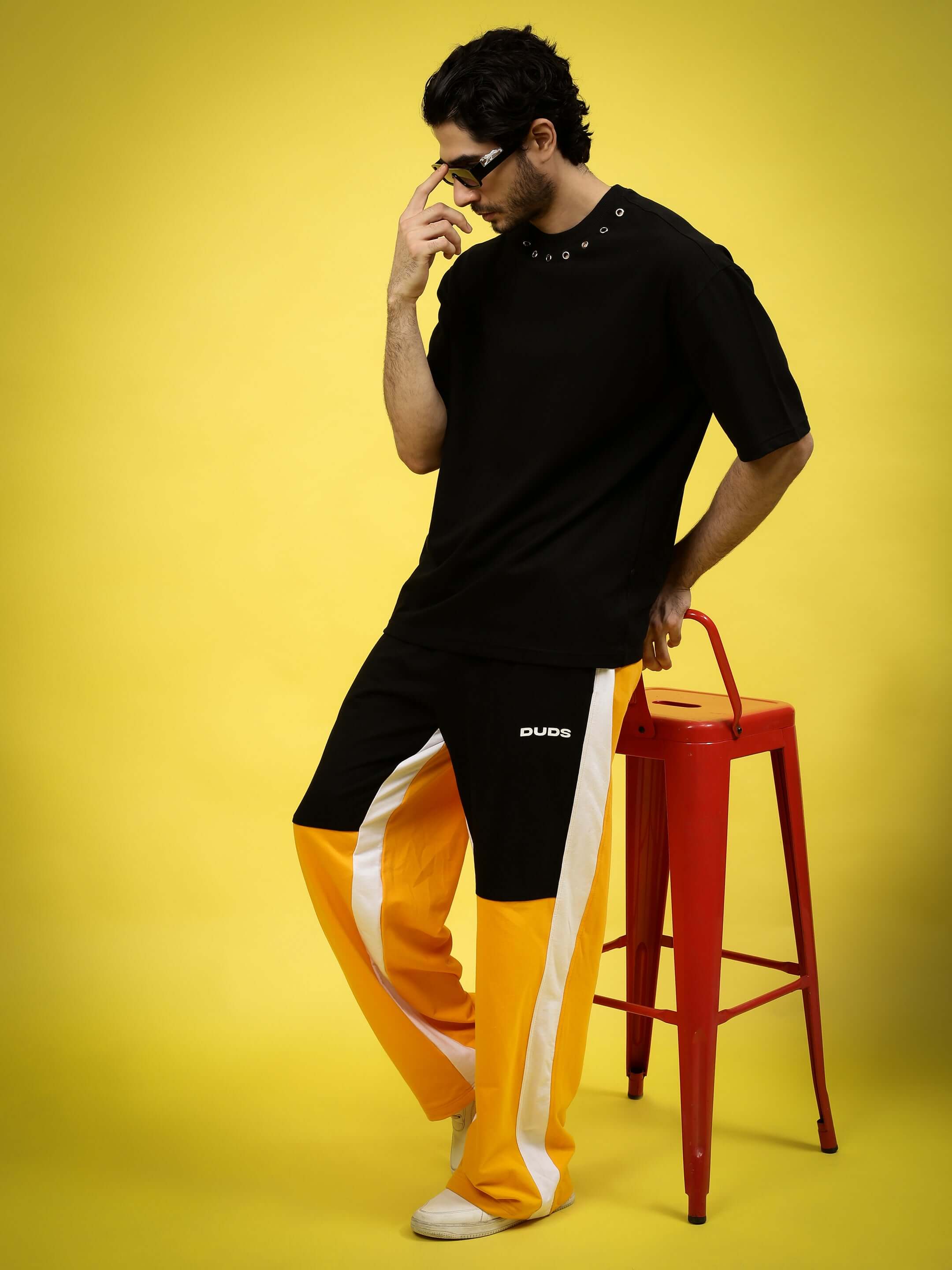 Man wearing PERILOUS OVER-SIZED T-SHIRT BLACK with bold graphic design, leaning on red stool against yellow background, showcasing edgy fashion style.