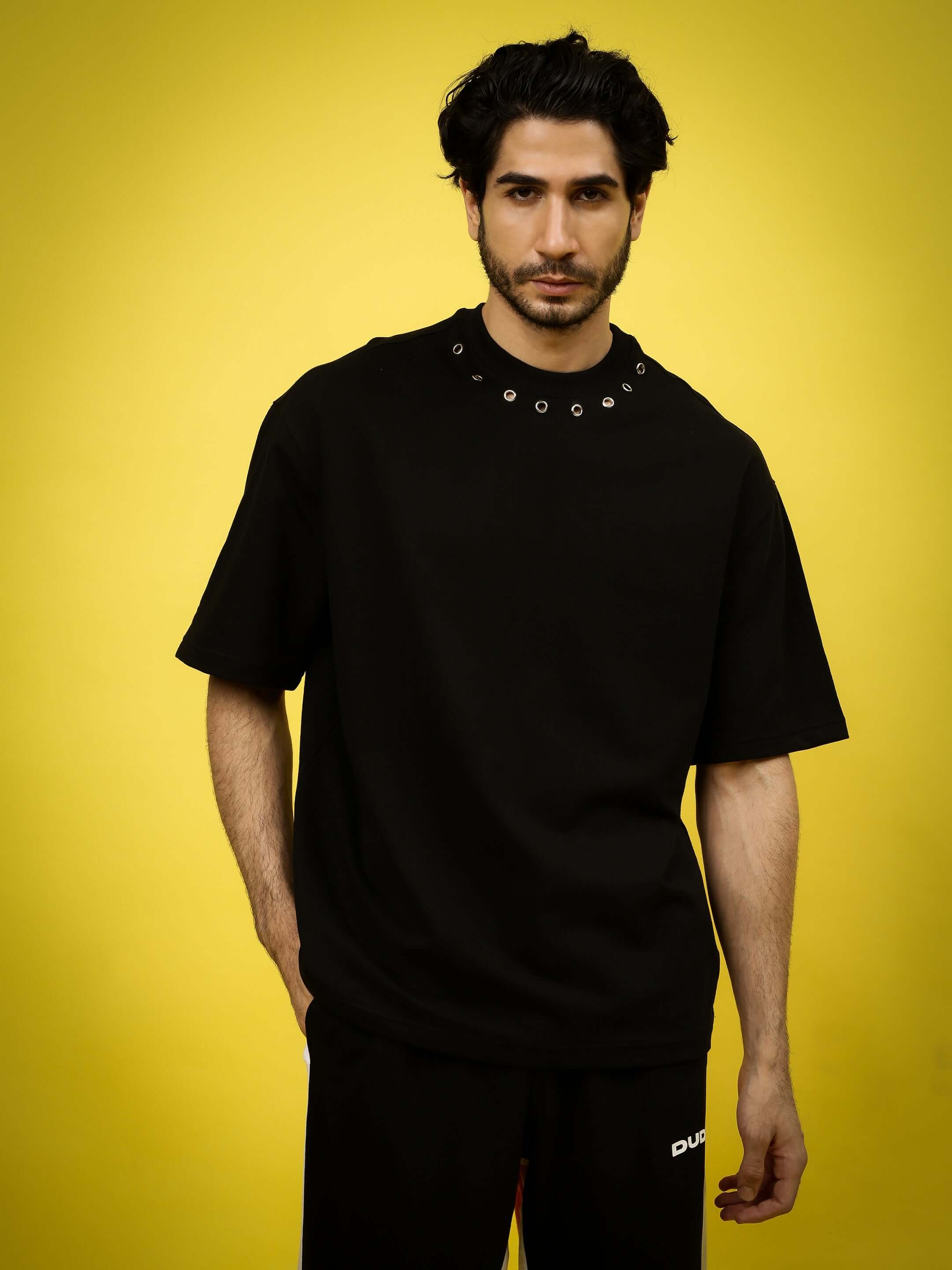 Man wearing a black Perilous oversized T-shirt with grommet details, standing against a yellow background, offered by DUDS.