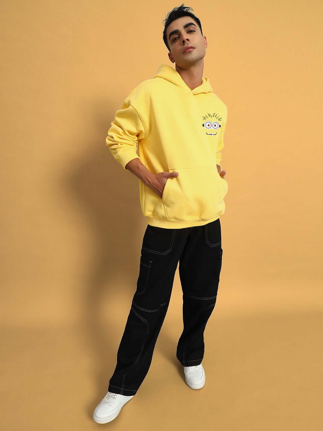Man wearing PHIL MINION OVERSIZED FLEECE HOODIE YELLOW paired with black pants and white sneakers, posing against a yellow background.