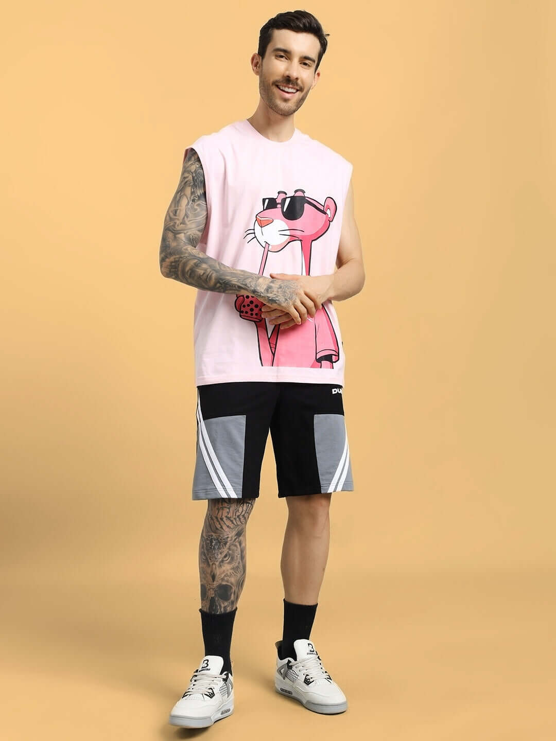 Man wearing casual outfit with graphic tank and patterned shorts, standing against a plain background.