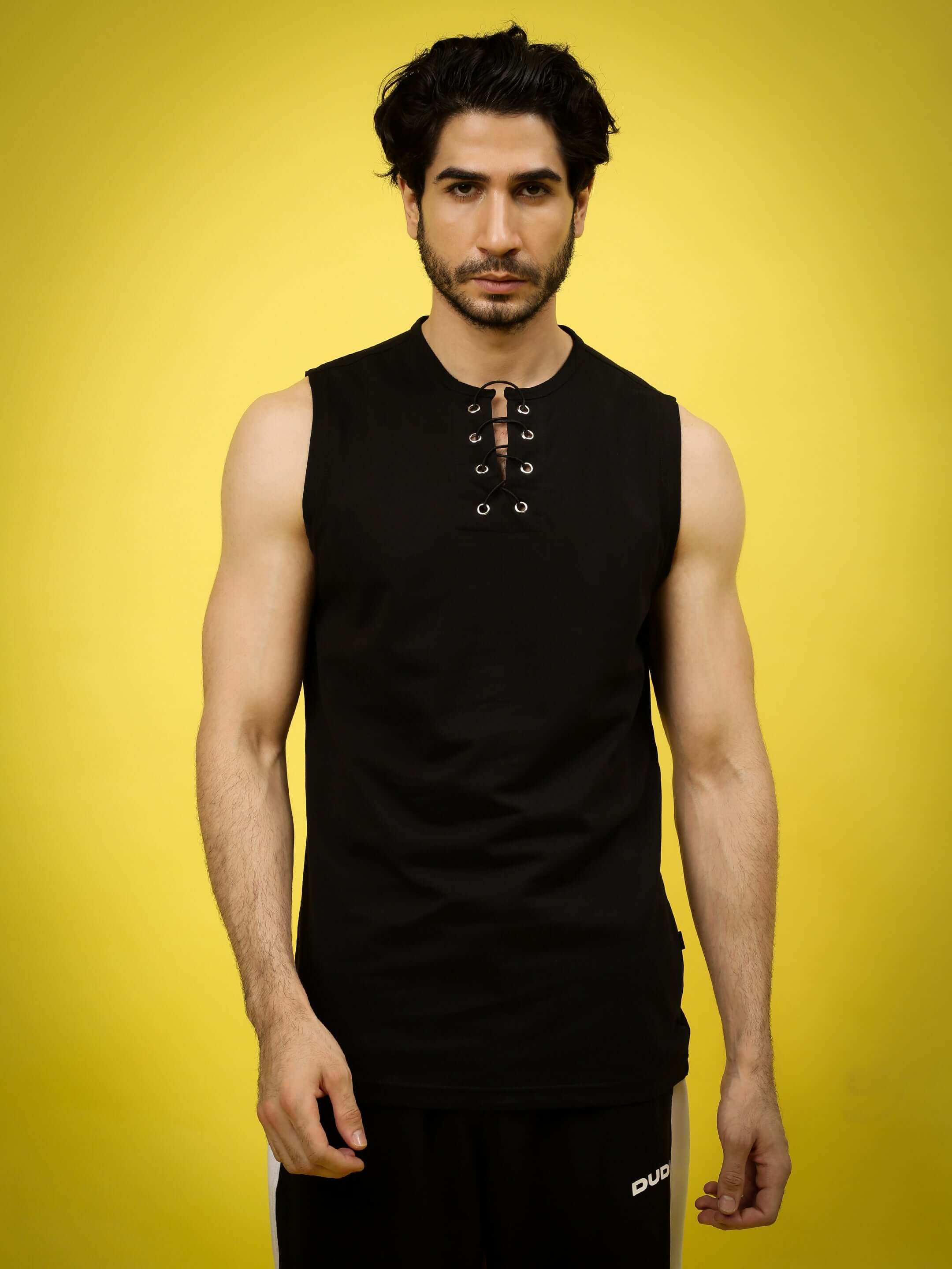 Man wearing a black sleeveless Pinnacle Regular Fit T-shirt with lace-up neckline, standing against a yellow background, showcasing contemporary style.