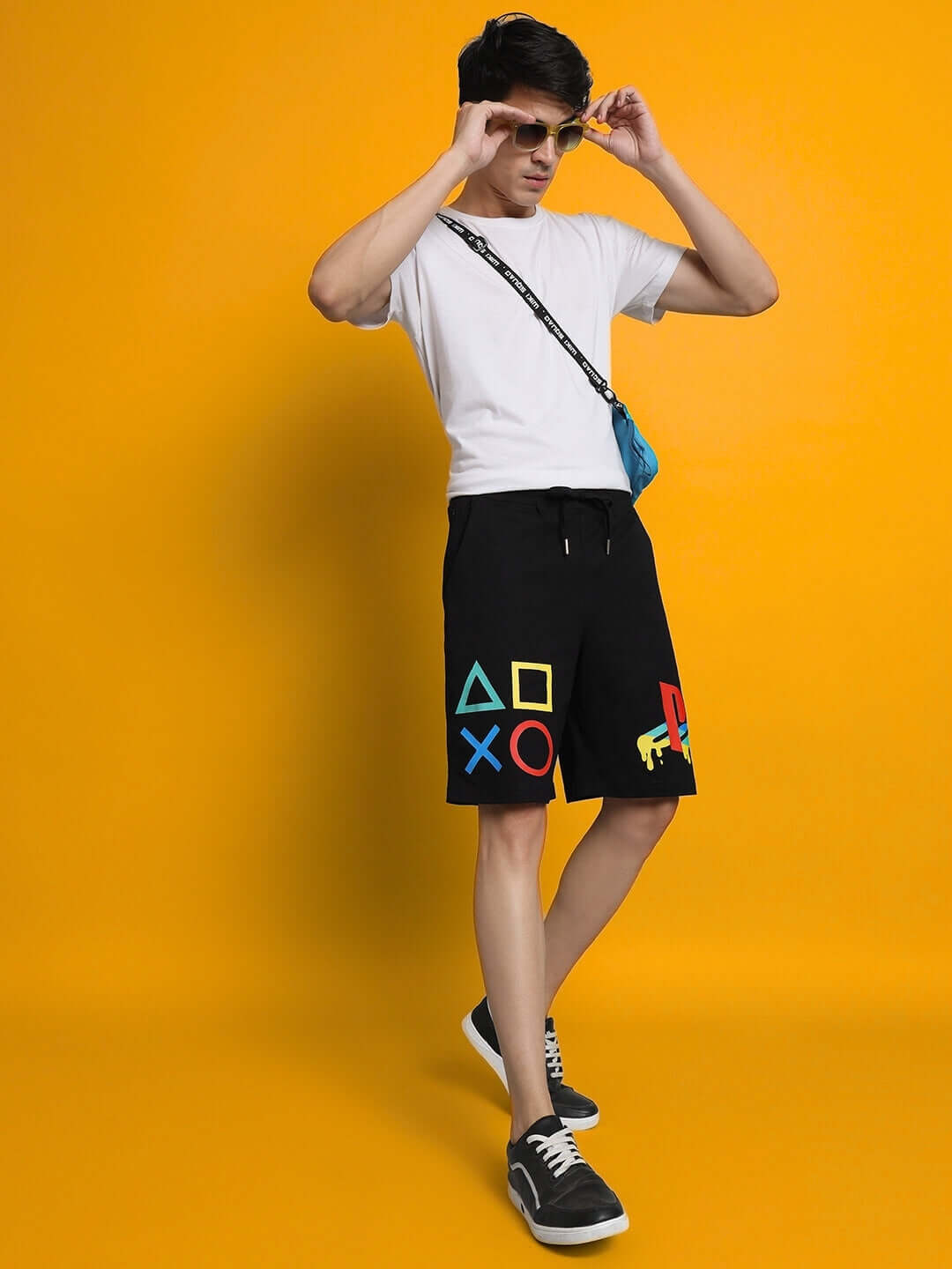Model wearing PLAYSTATION Regular Fit Black Shorts with iconic symbols, paired with a white tee and sunglasses on a yellow background.