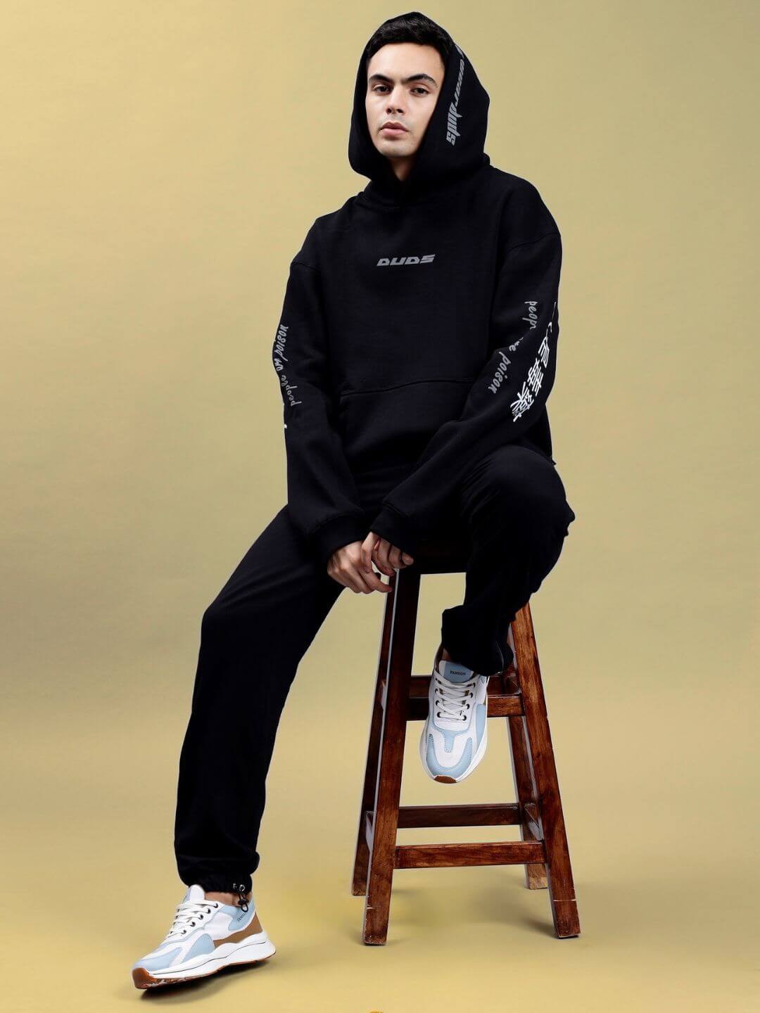 Person wearing POISON SKULL REFLECTOR OVERSIZE HOODIE BLACK sitting on a stool, showcasing streetwear style and reflective skull design.