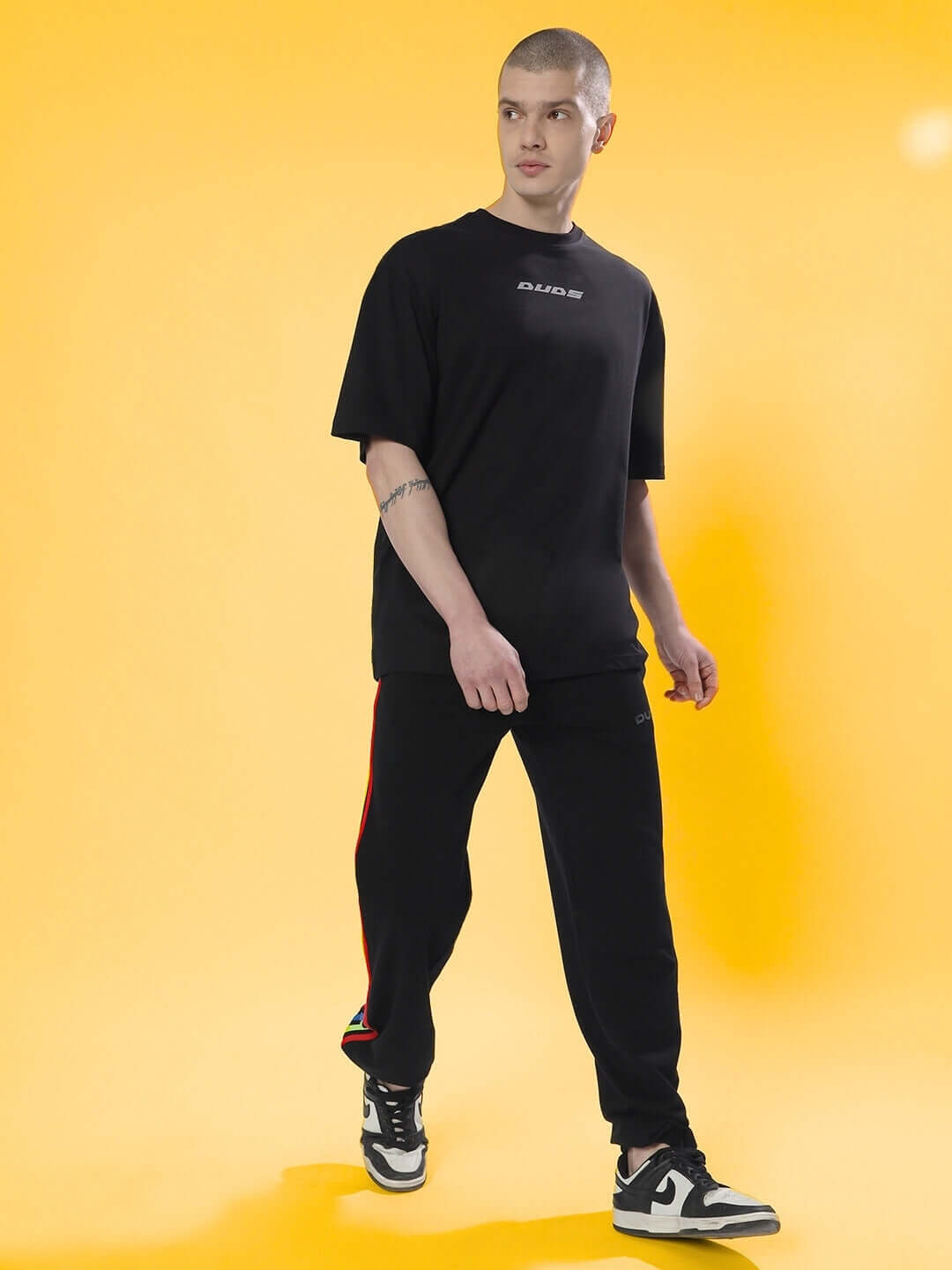 Model wearing oversized black T-shirt with skull design, paired with dark pants, on a yellow background. Casual fashion style.