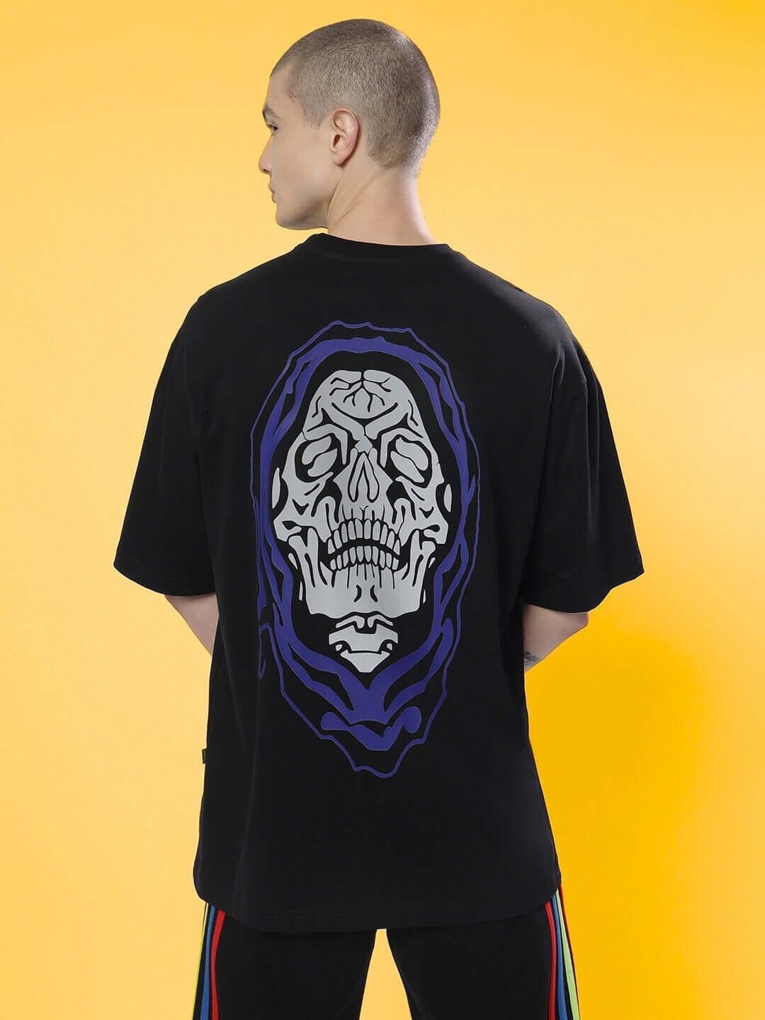 Man wearing black over-sized T-shirt with purple poison skull design on back against yellow background.
