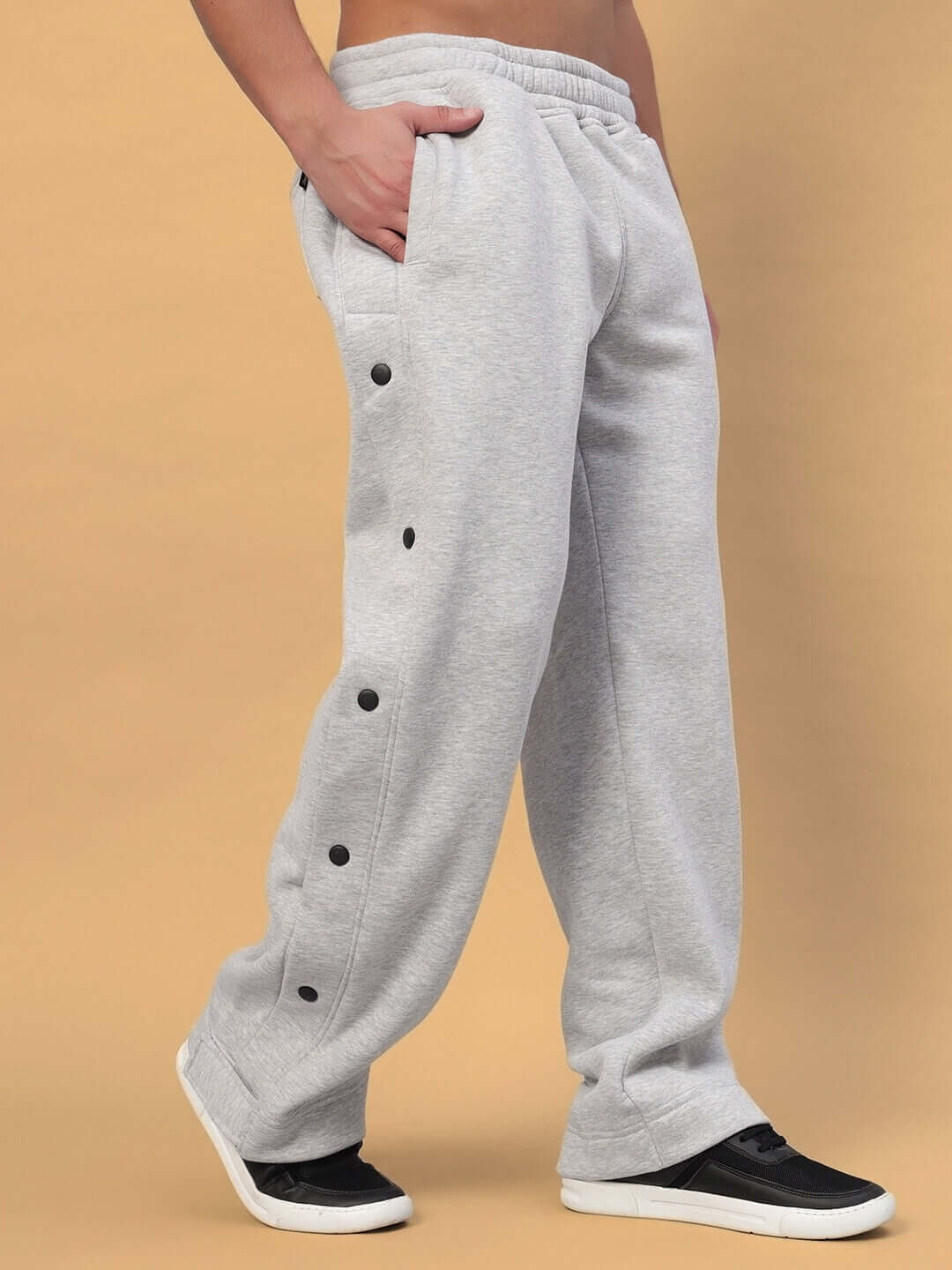 Person wearing grey POLAR cotton fleece jogger pants with black button accents and black sneakers, side view on beige background.