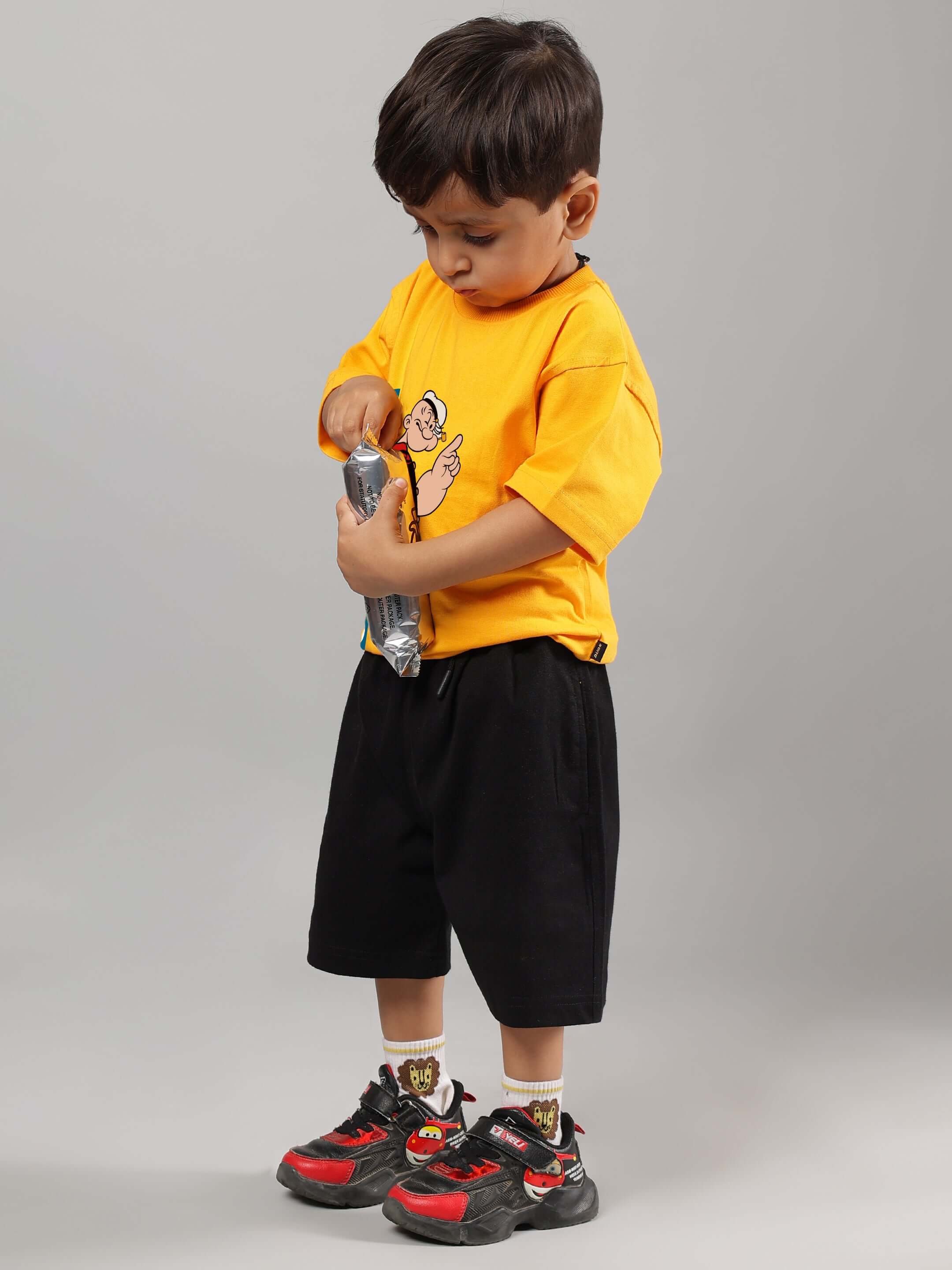 Child wearing a yellow Popeye t-shirt paired with black shorts and colorful sneakers, holding a water bottle.