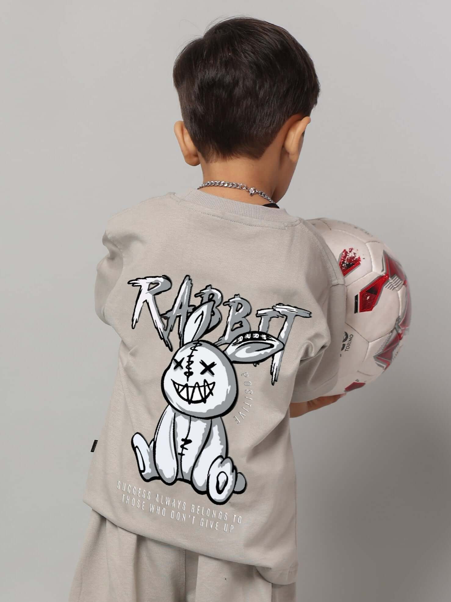 Child wearing grey Positive Rabbit T-shirt holding a soccer ball, showcasing the back graphic design.