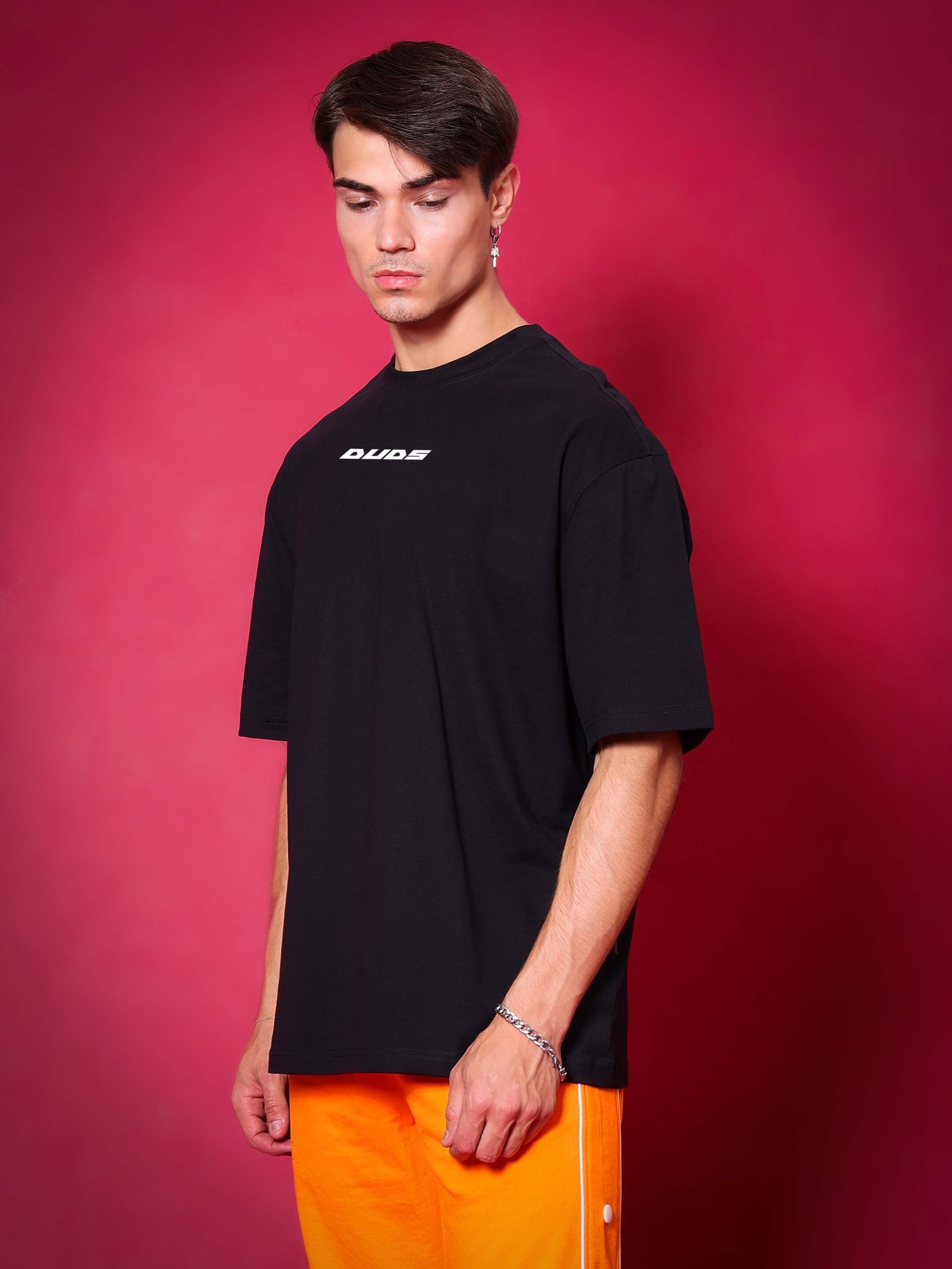 Model wearing a black oversized "PRAY" t-shirt made from premium cotton fabric, styled with vibrant orange pants against a pink background.