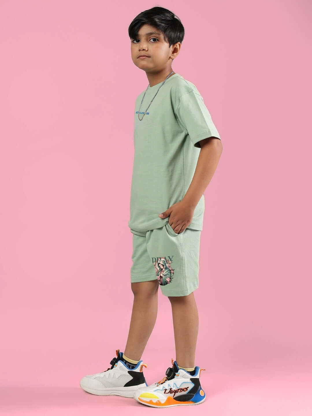Child wearing PRAY & PEACE CO-ORD FOR BOYS & GIRLS NYANZA GREEN outfit standing against pink background. Casual fashion style. Made to order clothing.