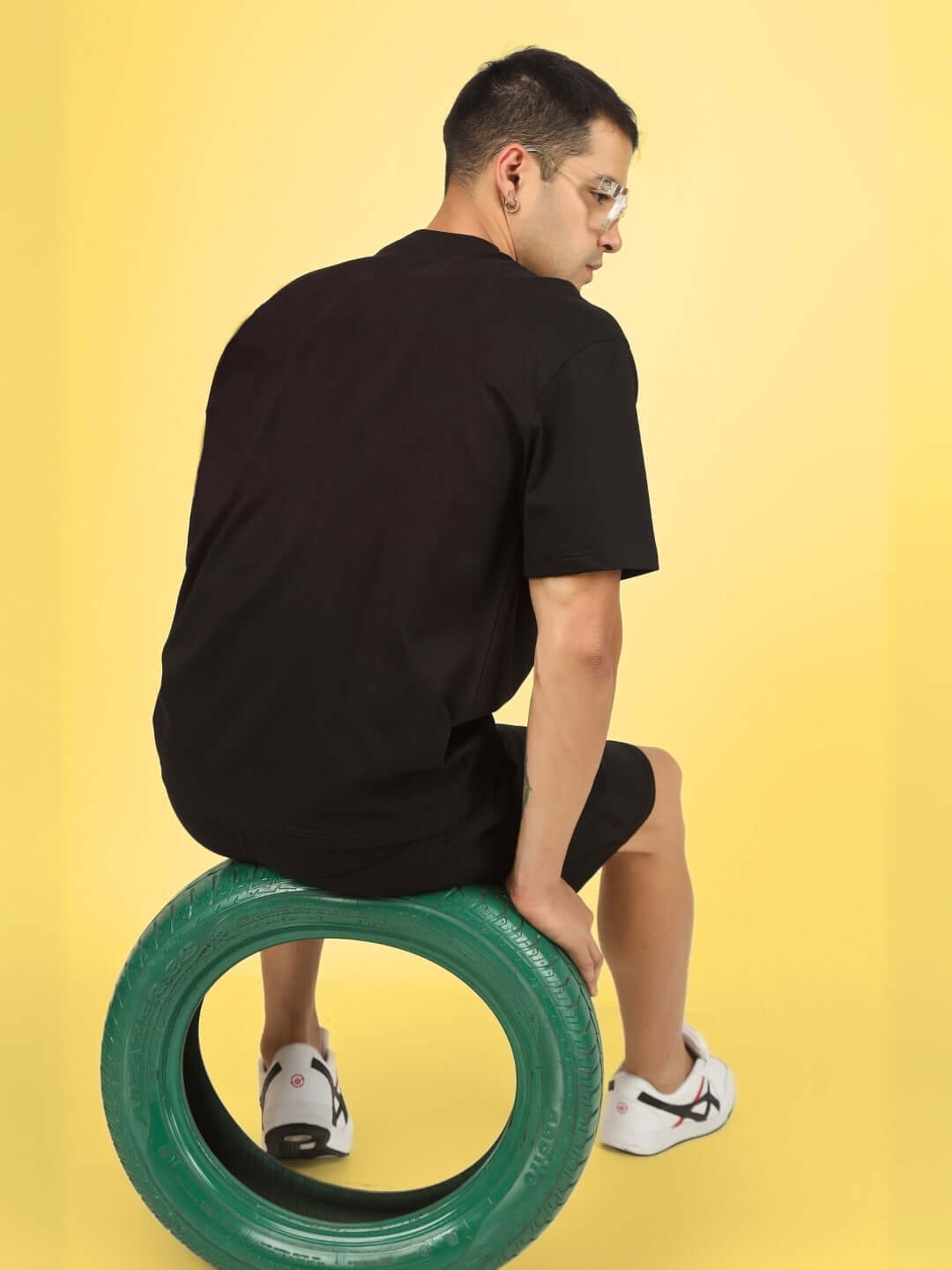Person wearing PREDATOR LEGND PRINTED SUMMER CO-ORD SET BLACK seated on a green tire with a yellow background.