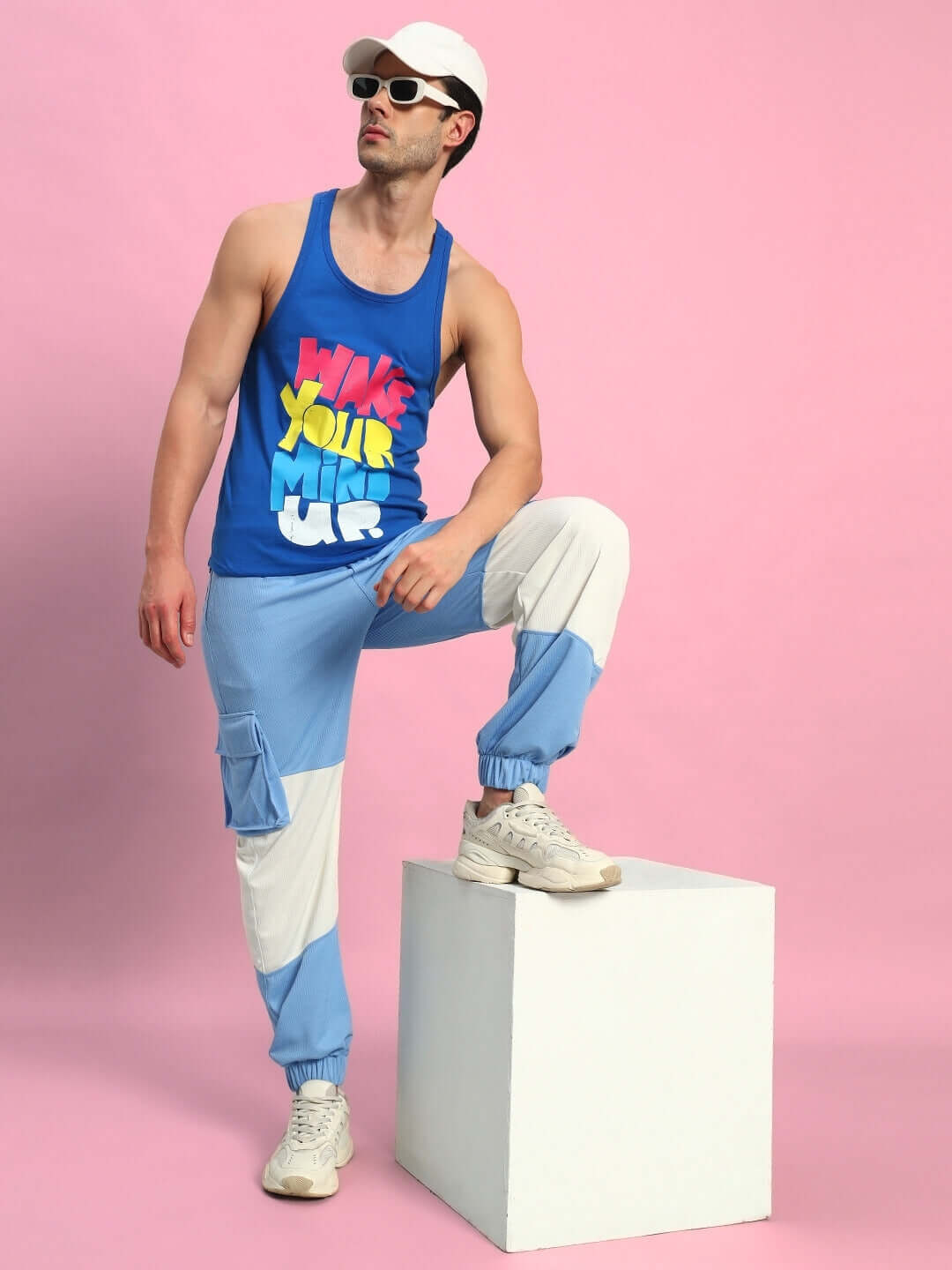 Man wearing Radiant Relaxed Fit Joggers in White-Sky Blue with casual tank top, standing on a white cube in front of pink background.