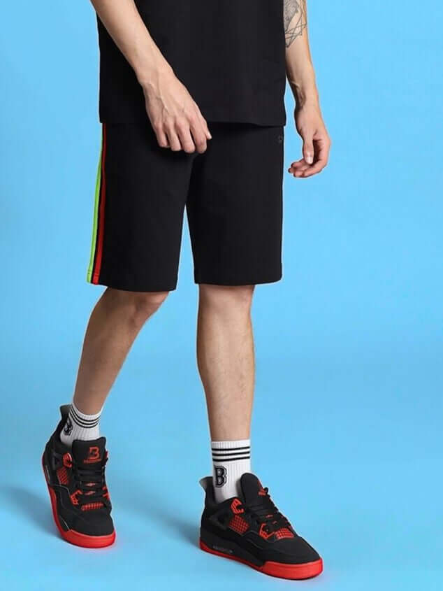 Man wearing RAINBOW REGULAR FIT SHORTS BLACK with stripe detail, standing on blue background, showcasing stylish, customizable fashion.