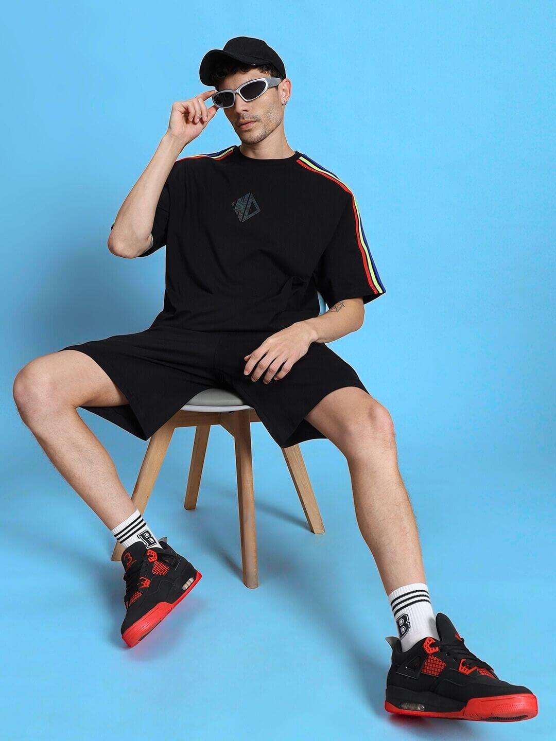 Model wearing Rainbow Summer Co-Ord Set in black with rainbow stripes, featuring premium cotton fabric and matching shorts, seated stylishly.