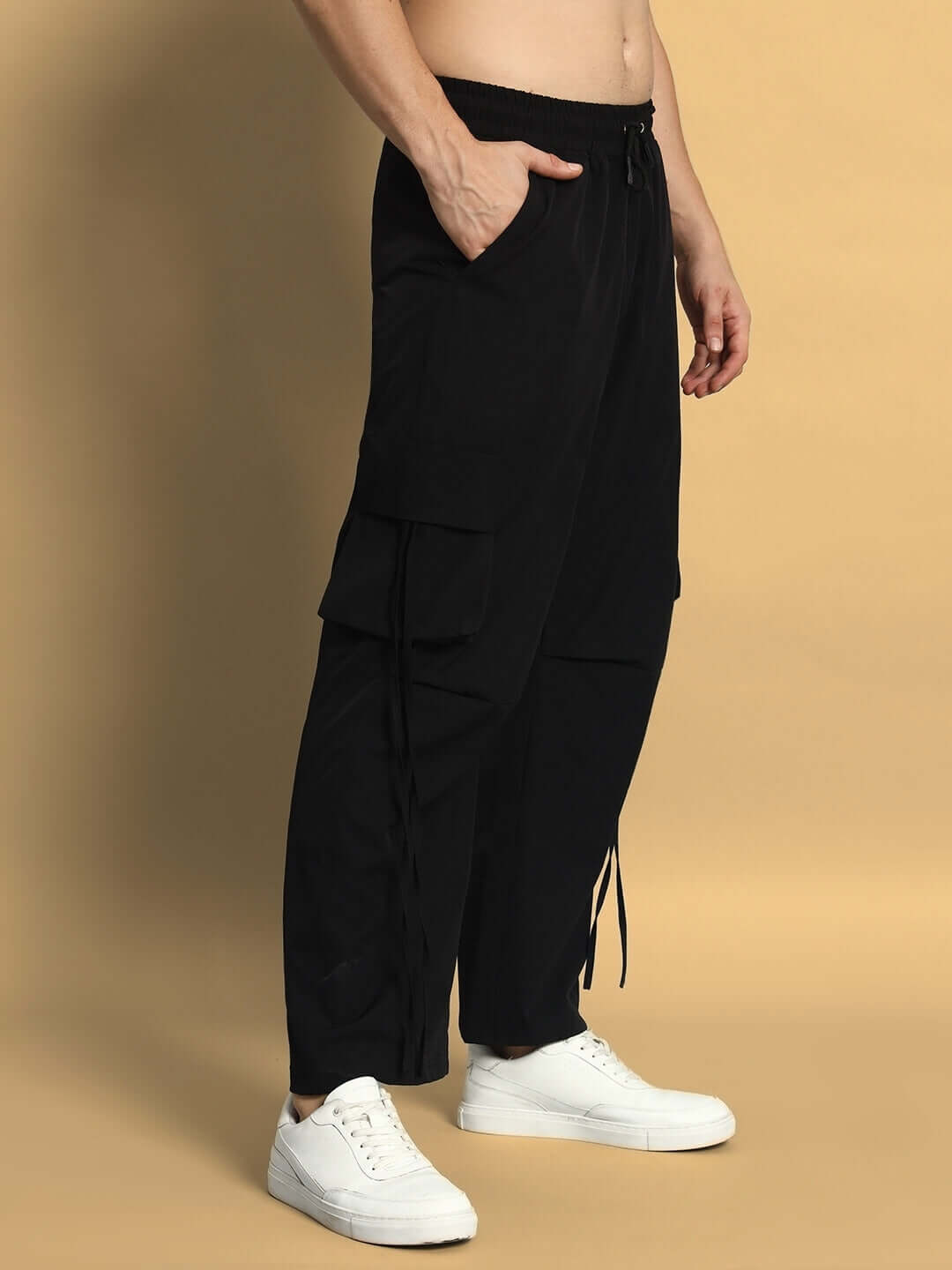 Model wearing black RAMROD parachute cargo pants with white sneakers, showcasing side pockets and drawstring details on beige background.