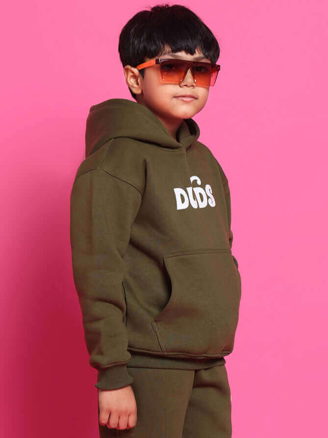 Child wearing olive green co-ord set with hoodie, posing in front of a pink background, wearing stylish sunglasses.