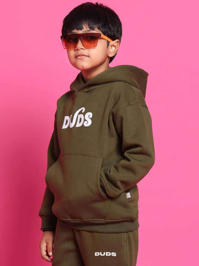 Child wearing olive green co-ord set with "DUDS" branding, stylish sunglasses, and hoodie against pink background.