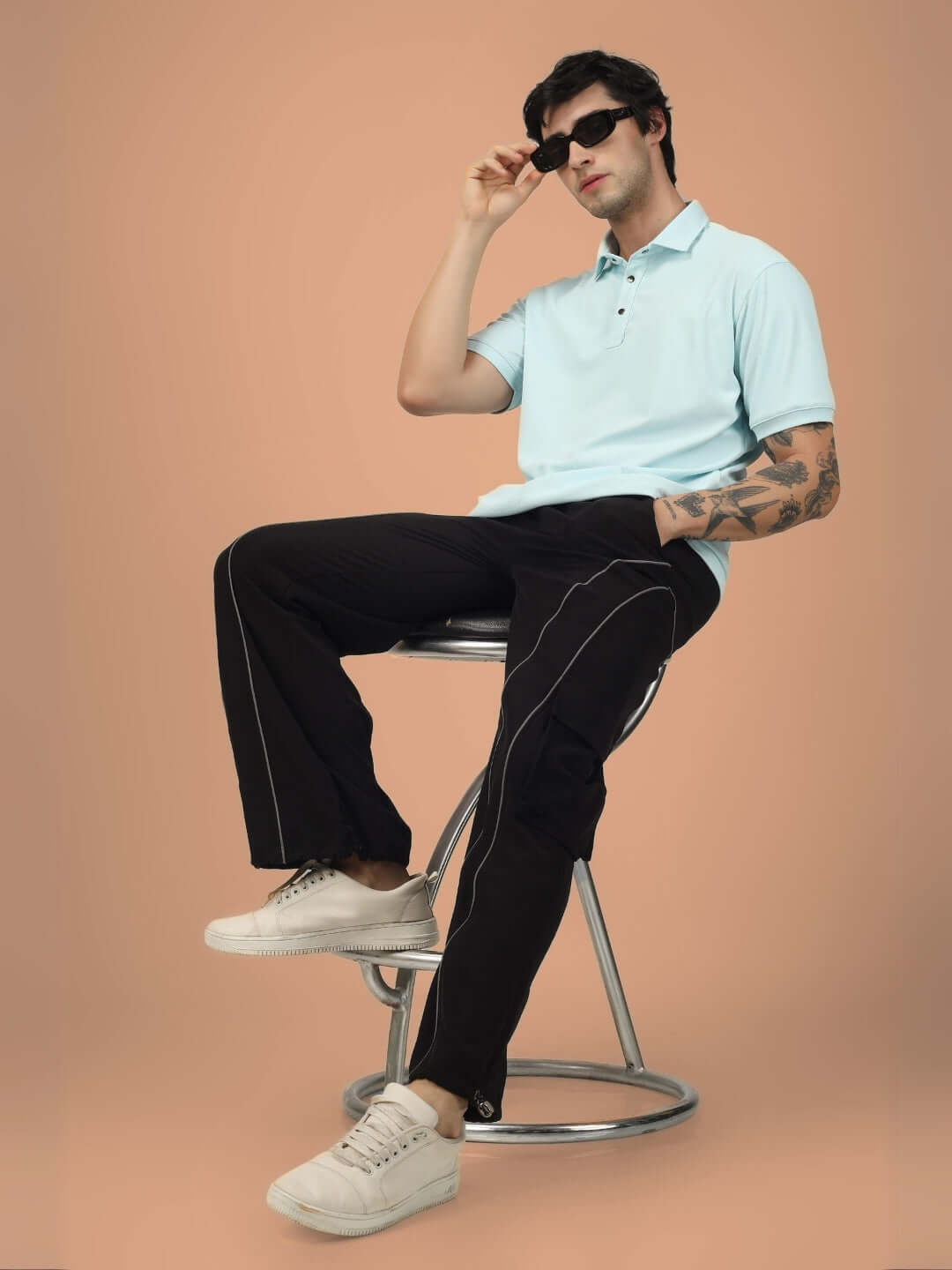 Model wearing Ray Reflector Parachute Cargo Pants in black, seated on a stool, showcasing casual style with a light blue polo shirt and sneakers.