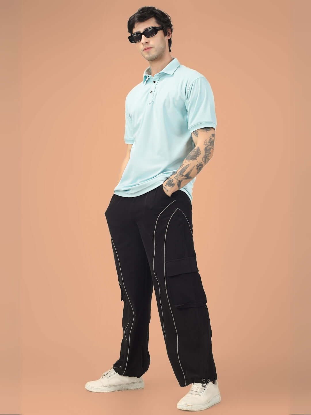 Man wearing Ray Reflector Parachute Cargo Pants in black with a light blue polo shirt and sunglasses on a peach background.