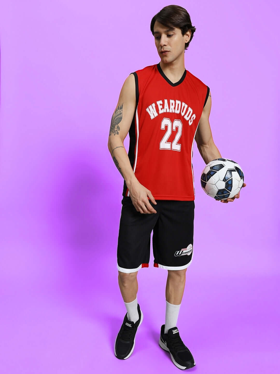 Man in red and black PORTIA gym co-ord set holding a soccer ball, standing against a purple background.