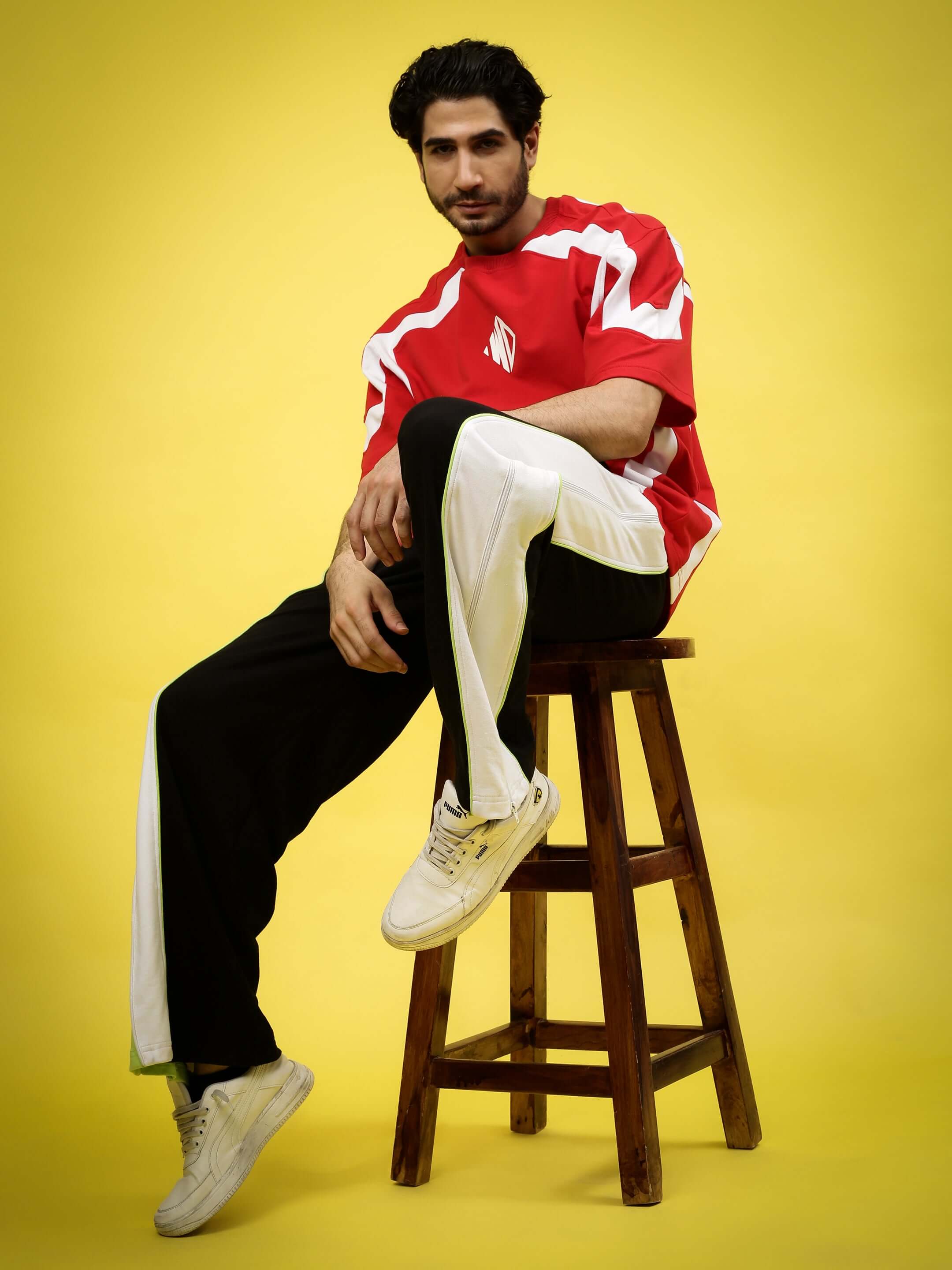 Man wearing a red sports shirt and black joggers seated on a wooden stool against a yellow background.