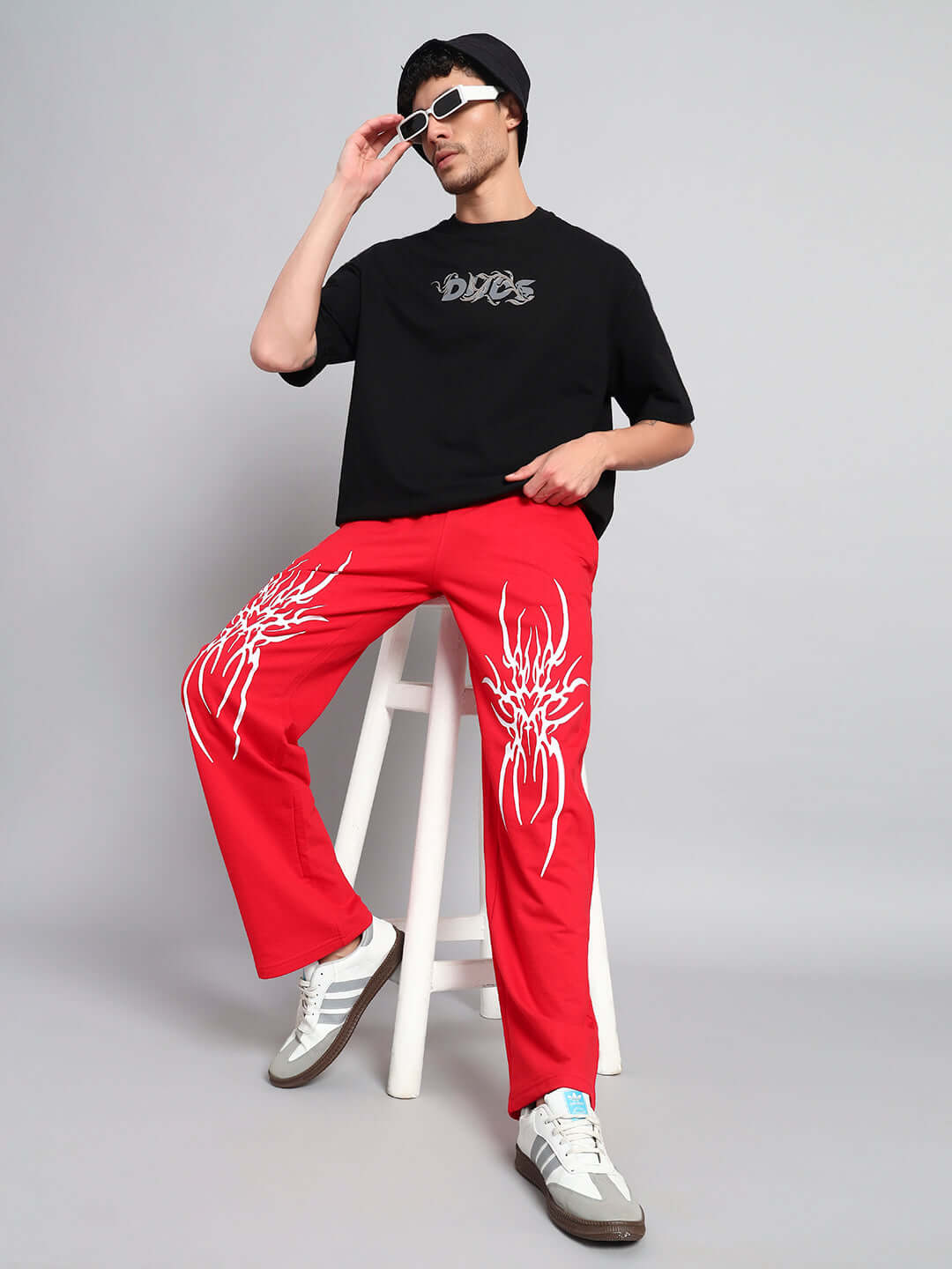 Model wearing REDX BRANCHING COTTON JOGGER RED MEN pants with bold graphics, sitting on a stool, showcasing trendy urban fashion.