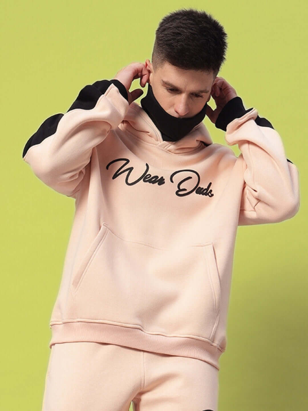Model wearing beige-black Regiment Fleece Hoodie with "Wear Duds" logo, highlighting comfort and style against green background.