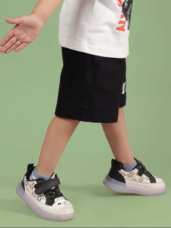 Child wearing ROBIN SHORTS FOR BOYS & GIRLS BLACK, walking in stylish sneakers against a green background.