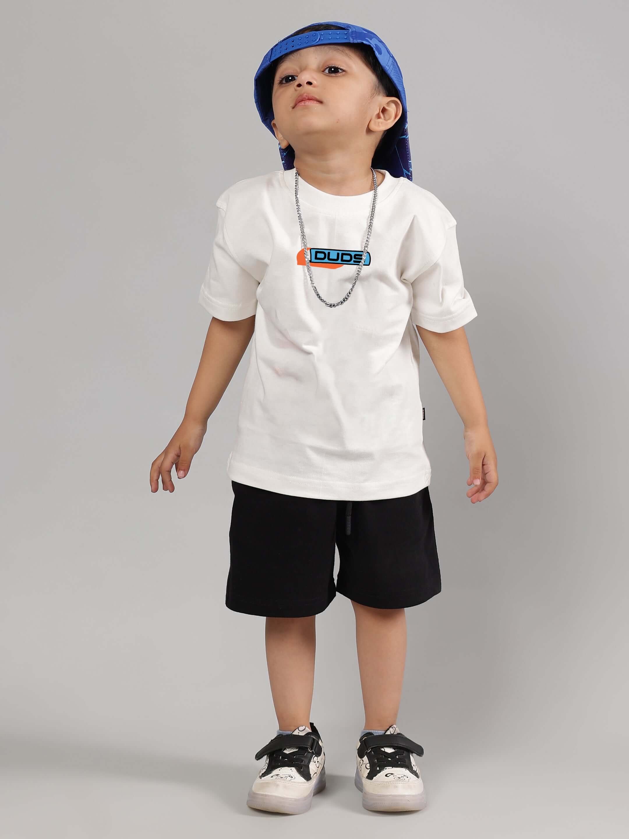 Child wearing DUDS white t-shirt and black shorts, part of the Robo Shark Co-ord for Boys & Girls, styled with blue hat and sneakers.