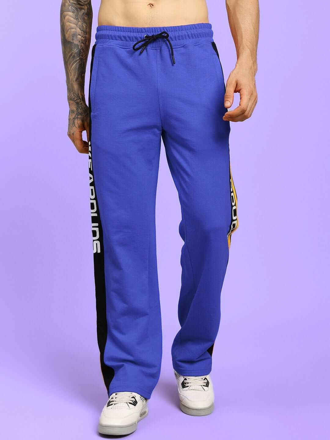 Man wearing Roger relaxed-fit blue joggers with side stripe, modeled with sneakers on a purple background.
