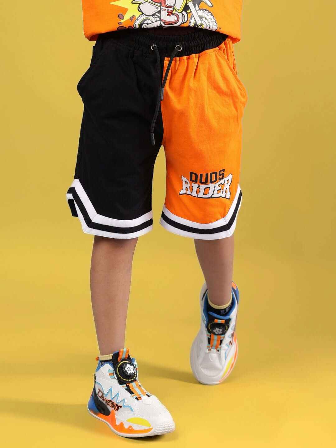 Child wearing black-orange Rogue Rider shorts, part of DUDS made-to-order collection, shown with colorful sneakers on yellow background.