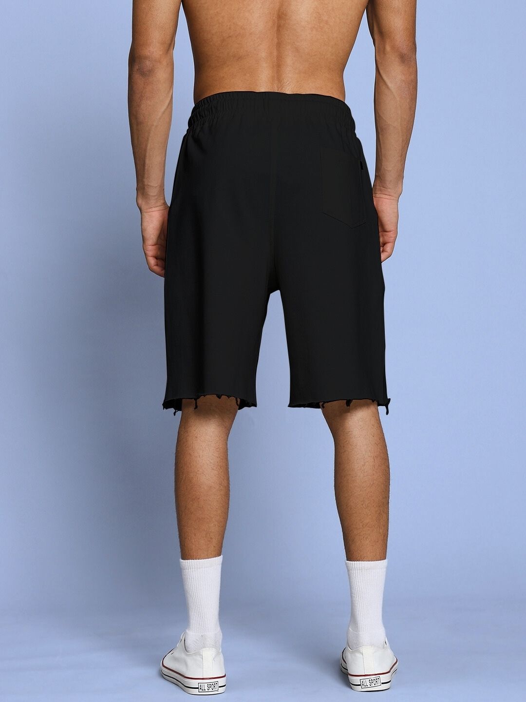 Back view of ROSE CHARM RELAXED FIT SHORTS BLACK shown on a male model standing against a blue background, featuring a casual style.