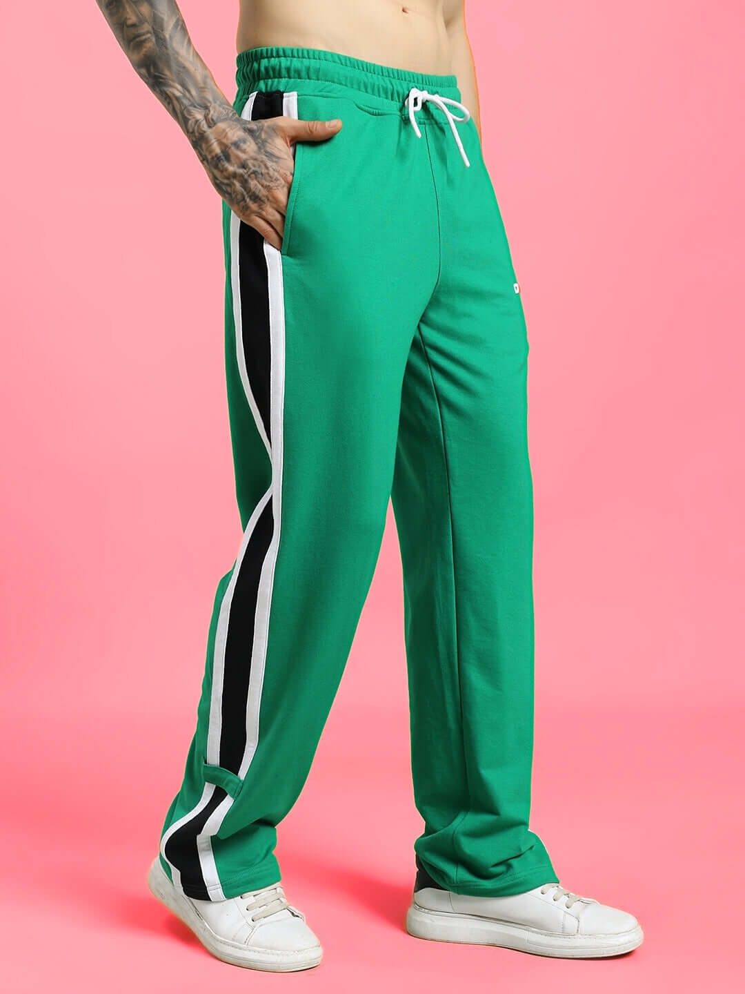 Model wearing ROSSTON FLAT-OUT RELAXED FIT JOGGERS GREEN with side stripes and white sneakers on pink background.