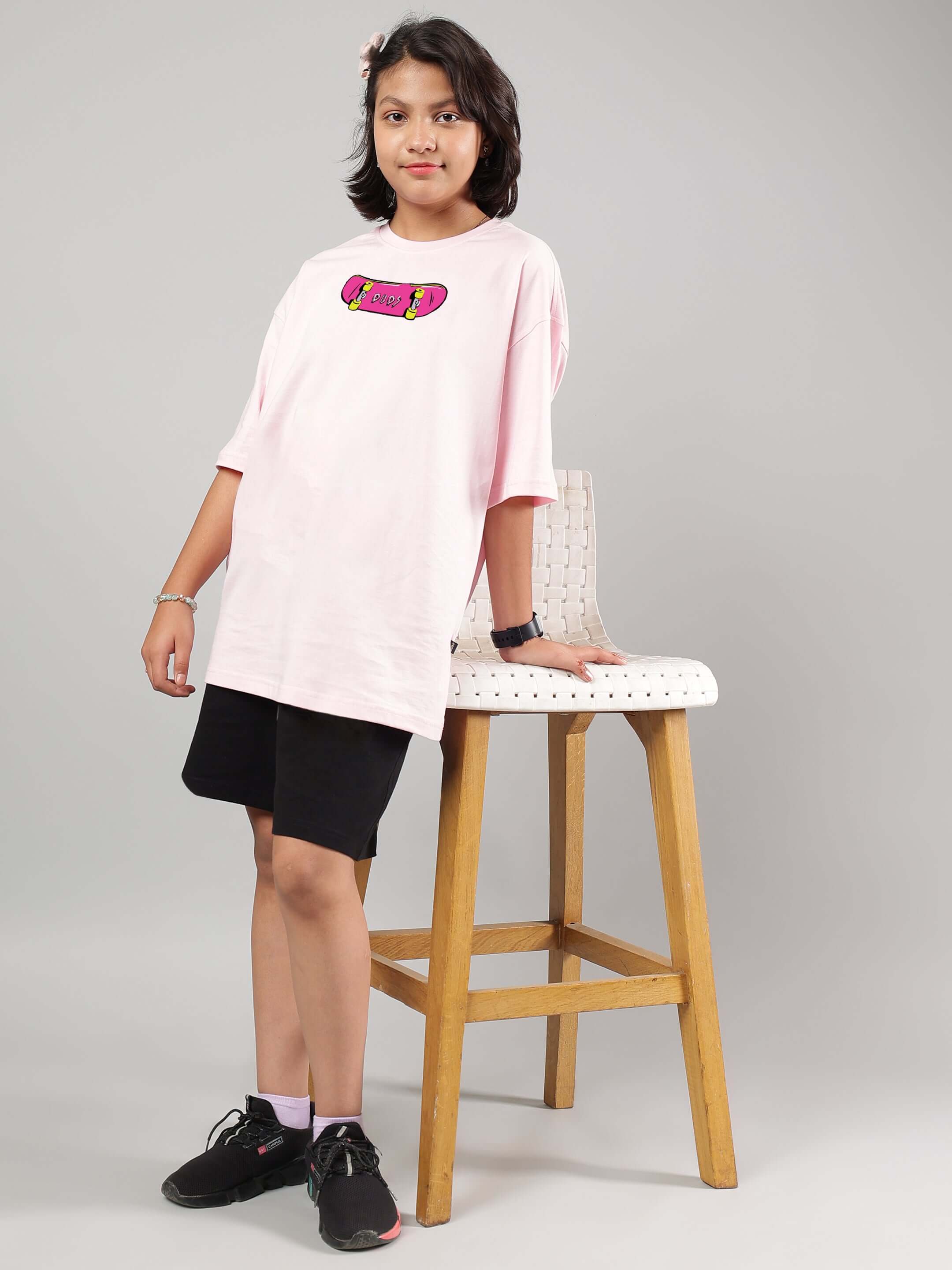 Child wearing a pink oversized t-shirt for boys and girls, featuring a graphic design, standing next to a tall wooden stool.