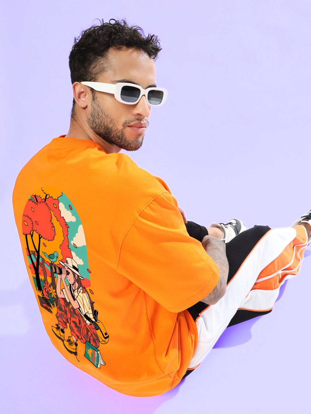 Man wearing SAFARI PRINTED OVERSIZED T-SHIRT ORANGE with graphic design, sitting against a lavender background.
