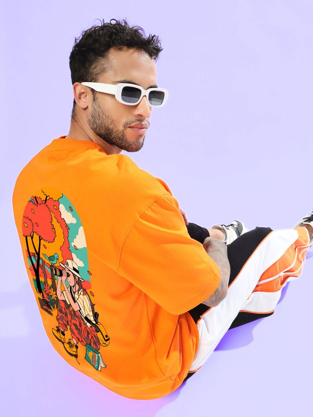 Man wearing a vibrant SAFARI PRINTED OVERSIZED T-SHIRT ORANGE with a bold safari print, sitting casually against a purple background.