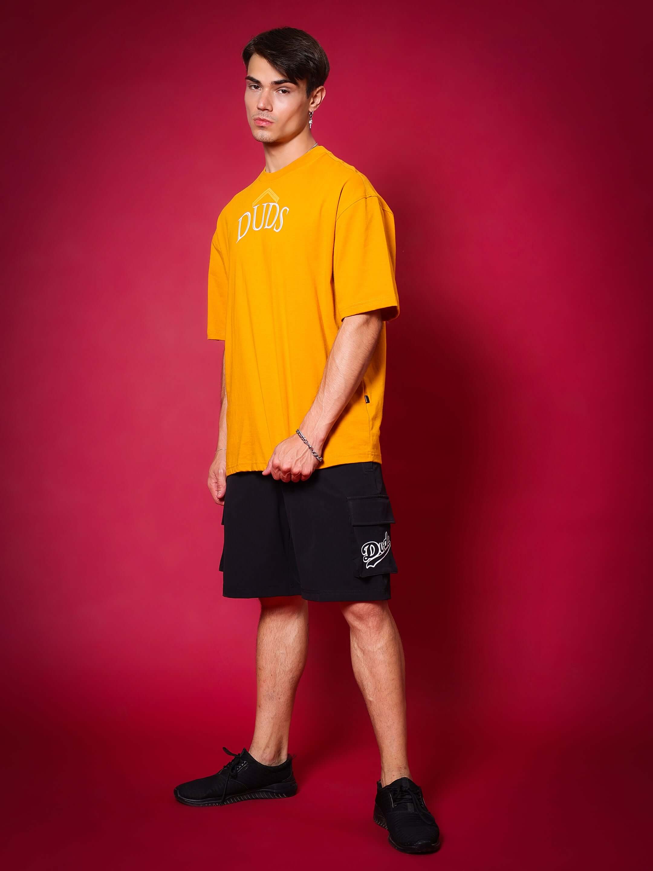 Model wearing SAKURA Blossom over-sized mustard T-shirt by DUDS, styled with black shorts, standing against a red background.