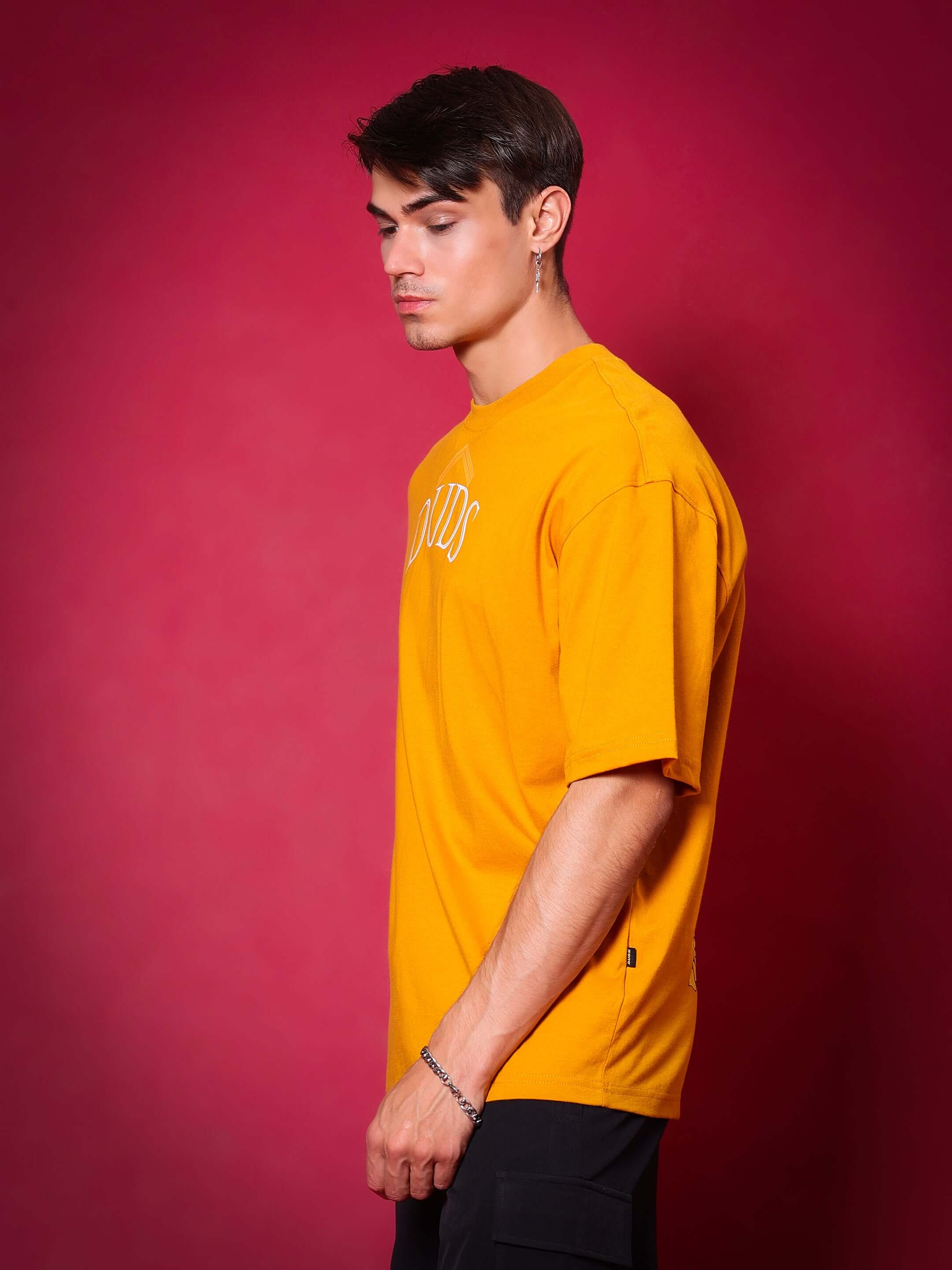 Model wearing mustard SAKURA BLOSSOM over-sized T-shirt with DUDS logo, standing against a red background.